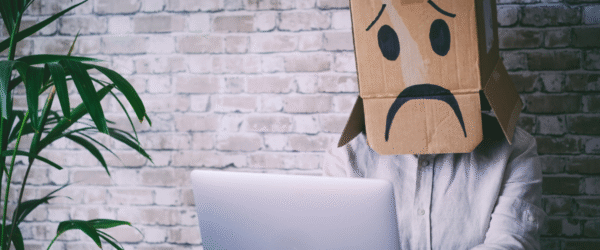 A person sitting in front of a compute with a cardboard box on their head with a sad stressed face drawn on it.